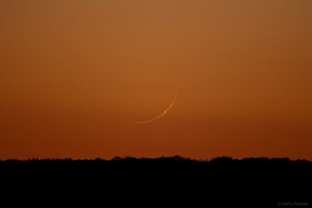 Vrlo tanak Mjesečev srp, malo izobličen atmosferskom distorzijom, na crvenom nebu sumraka