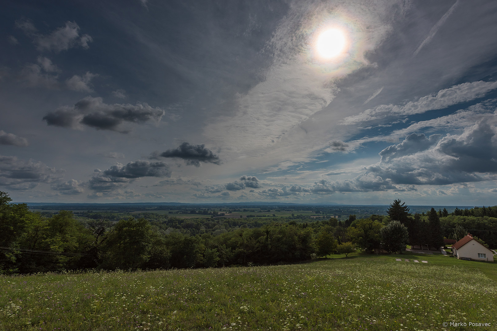 Pogled na ravnicu s travnatog brda, na nebu je više vrsta oblaka, a oko Sunca se vidi niz raznobojnih prstenova