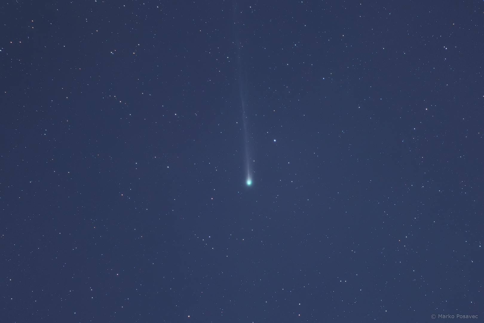 Komet sa zelenkastom jezgrom i blijedim, tankim repom usmjerenim vertikalno gore na pozadini lagano plavičastog zvjezdanog neba