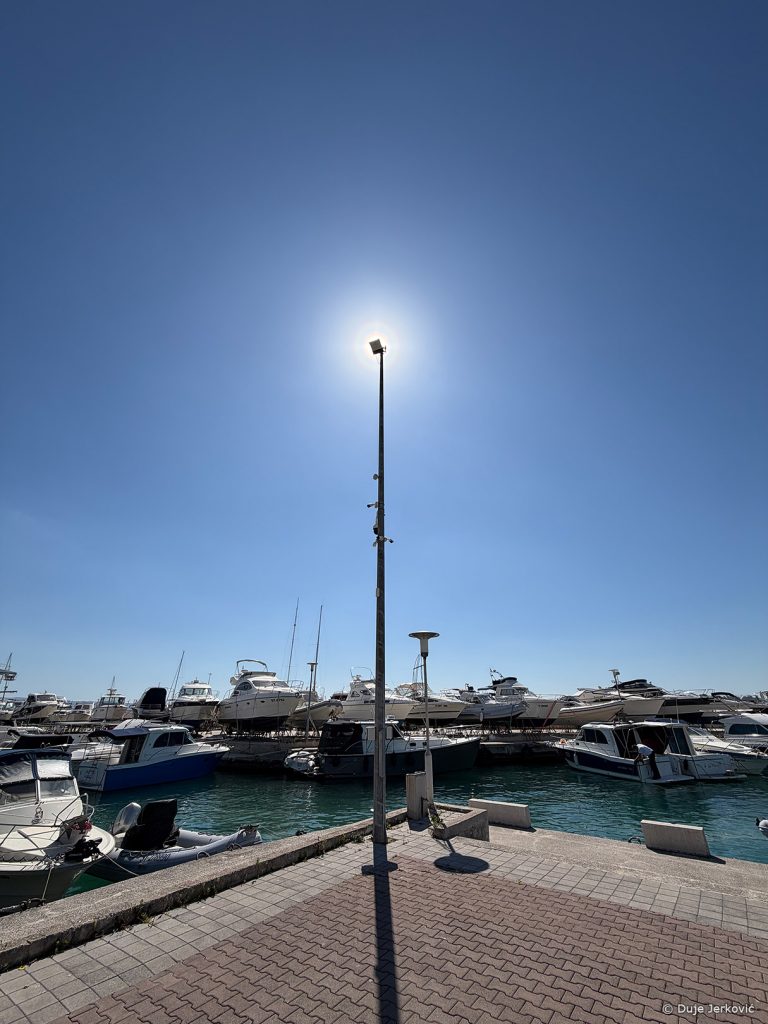 Pogled na lučicu s brodicama, u sredini slike je rasvjetni stup koji zaklanja Sunce, a oko njega se vidi peludni vijenac